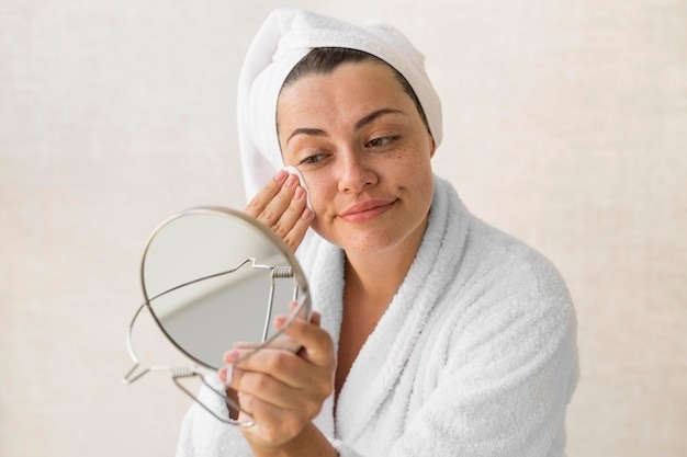 Mulher de toalha na cabeça e roupão, colocando em prática sua rotina de skincare para pele oleosa em frente ao espelho.
